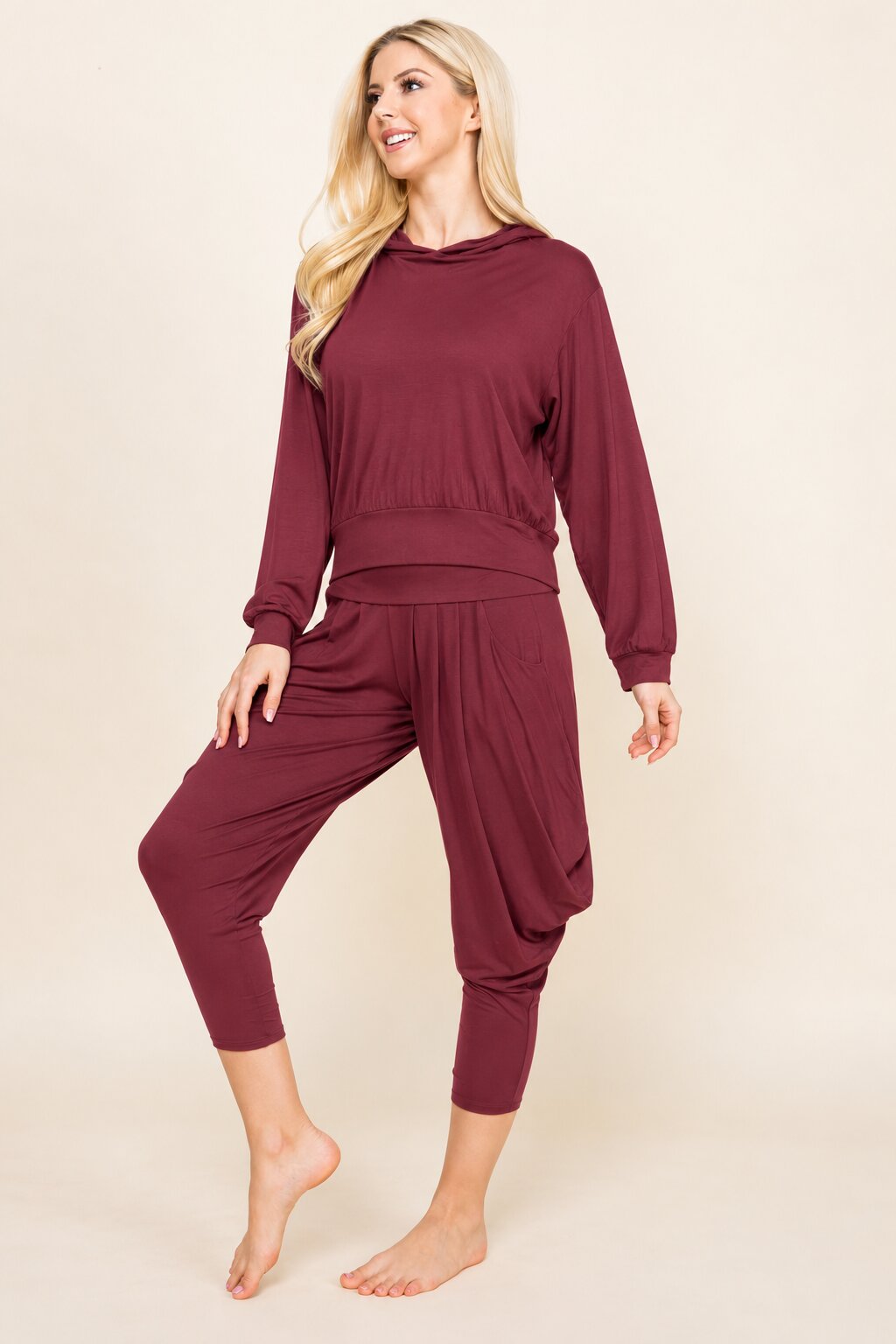 A woman modeling a matching burgundy lounge set consisting of a long-sleeve cropped sweatshirt and matching jogger-style pants, photographed against a solid beige studio background.