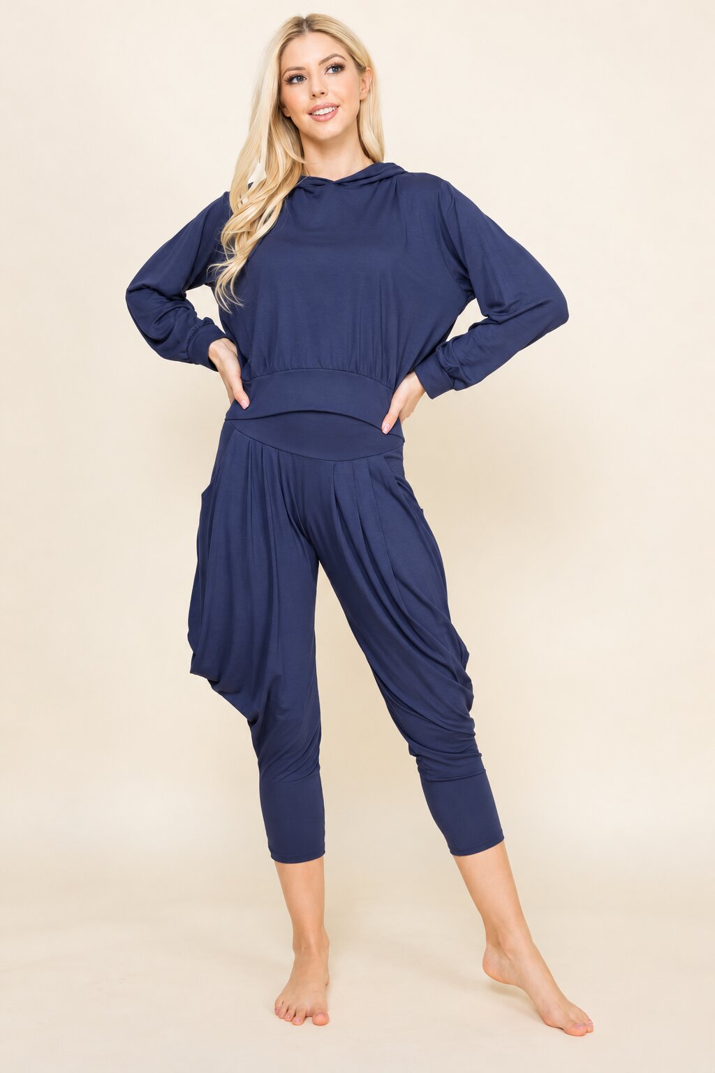 A woman modeling a navy blue two-piece loungewear set consisting of a long-sleeve cropped hoodie and matching draped capri pants, posed against a solid beige studio background.