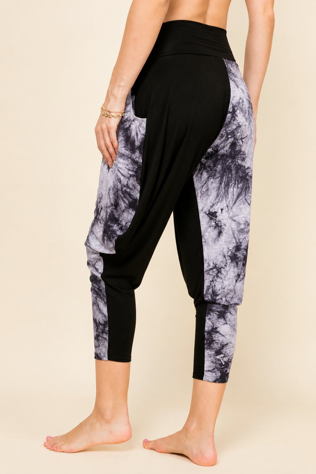 A person wearing high-waisted cropped harem-style leggings with black side panels and gray-and-white tie-dye side panels, shown from the back against a plain beige studio background.