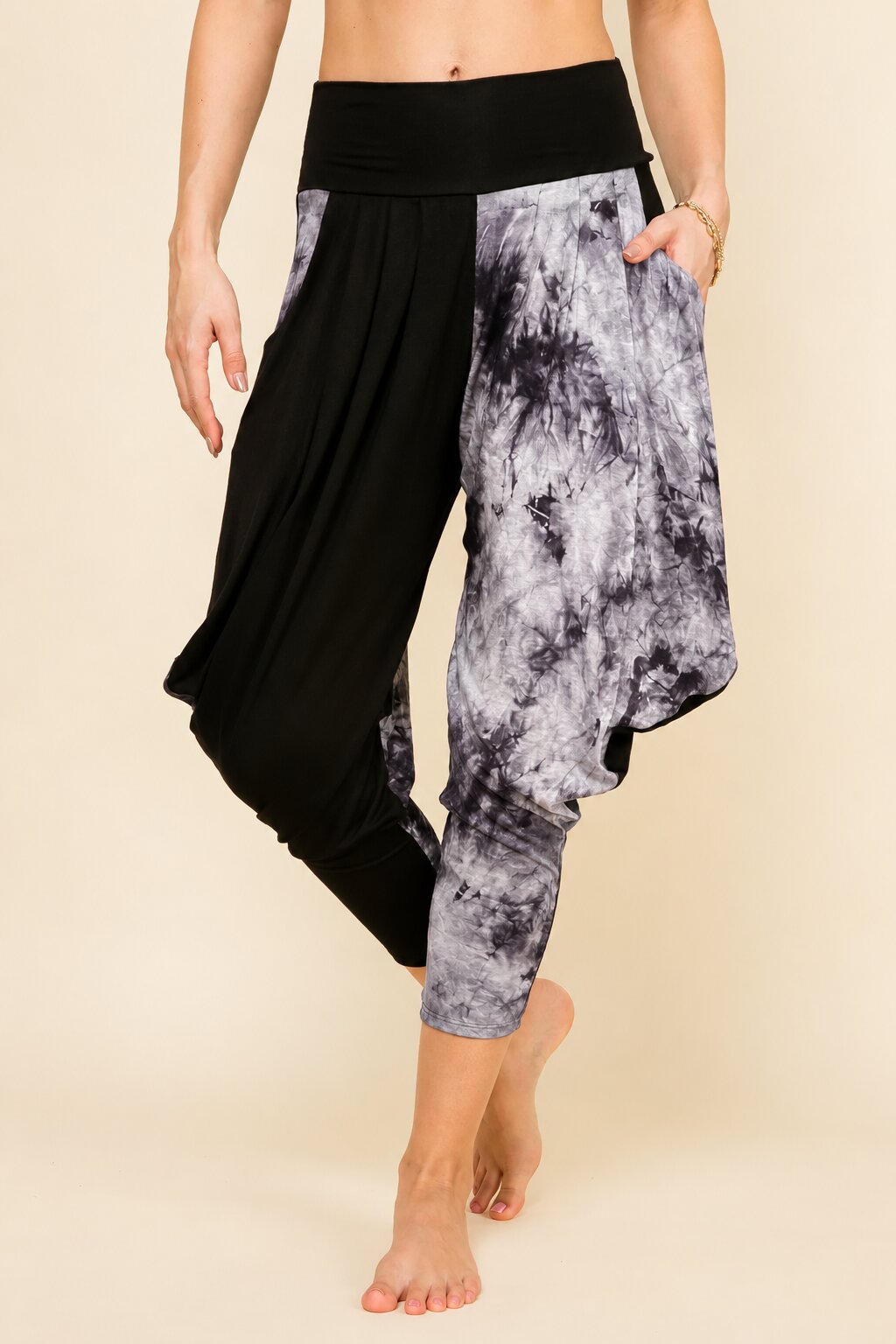 A person modeling loose harem-style pants with a black left leg and a gray, white, and black marbled print right leg, against a plain beige studio background.