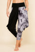A person modeling loose harem-style pants with a black left leg and a gray, white, and black marbled print right leg, against a plain beige studio background.