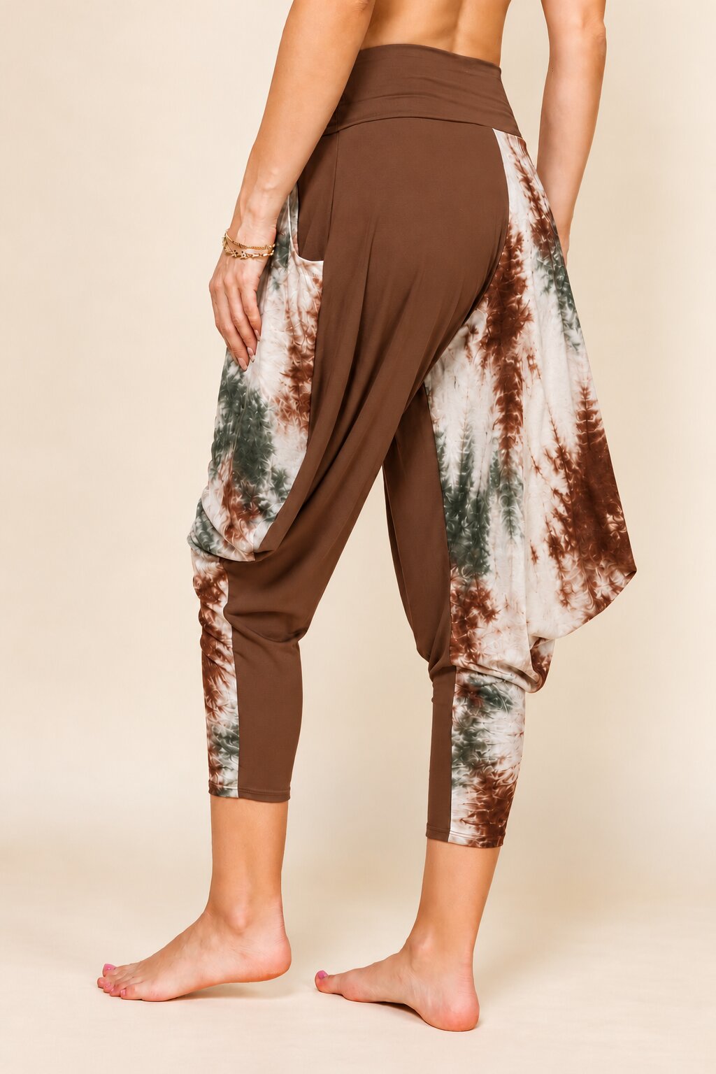 A person wearing brown harem-style capri leggings with side tie-dye panels in white, brown, and green, shown from the back against a plain beige studio background.