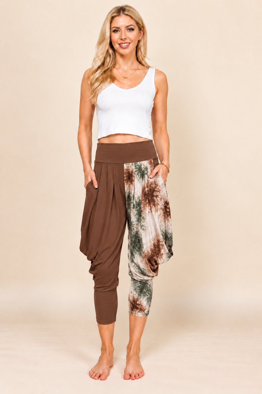 A woman modeling high-waisted harem-style cropped pants with one solid brown leg and one side-slit patterned leg in brown, green, and cream tie-dye, paired with a white crop top against a beige studio background.