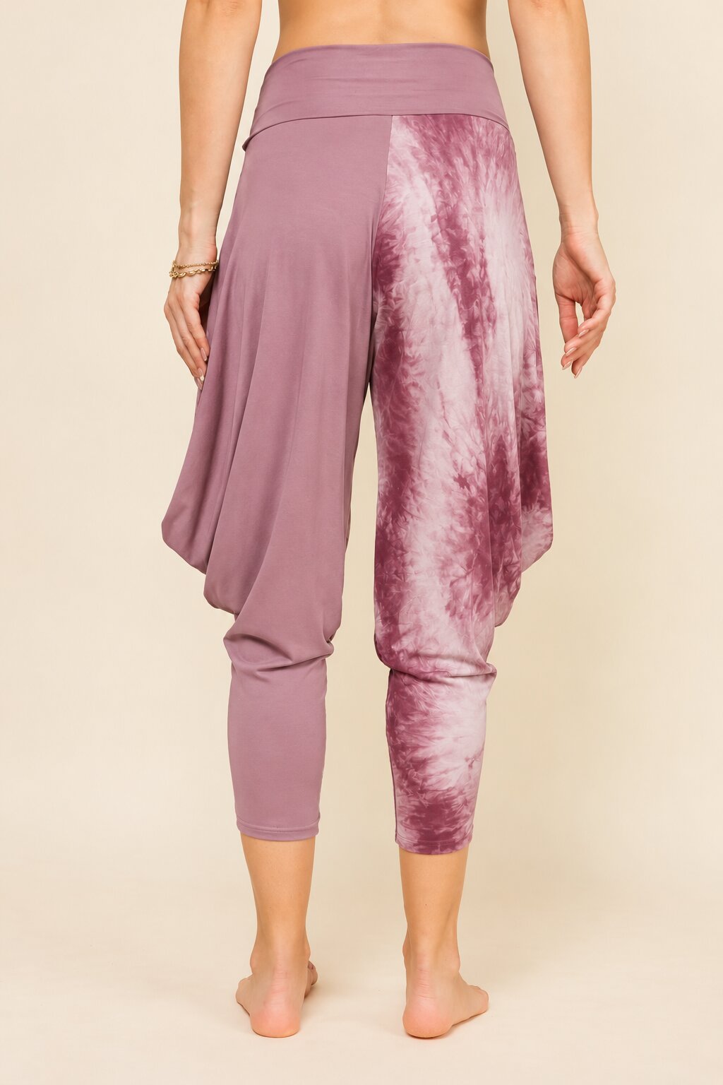 A person shown from the back wearing loose, draped capri leggings with a purple and pink tie-dye pattern, standing against a plain beige studio background.