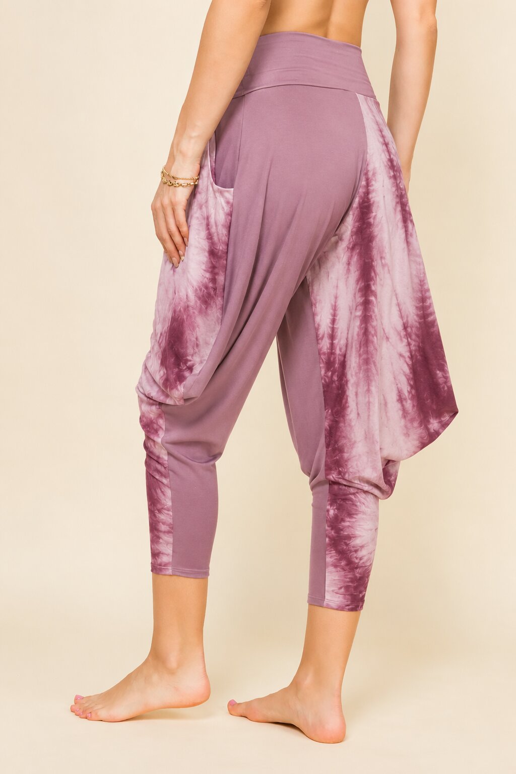 A model from the back wearing dusty purple harem-style capri leggings with a white tie-dye pattern, standing barefoot against a solid beige background.