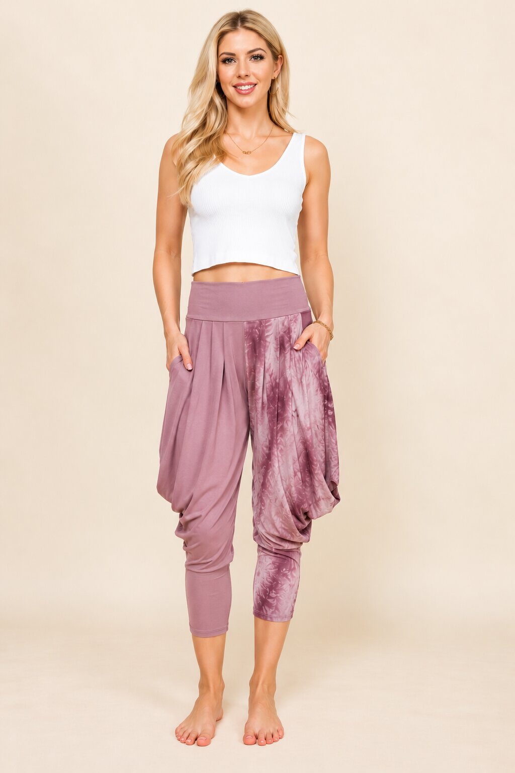 A woman modeling dusty purple harem-style cropped pants with a tie-dye pattern on one leg, paired with a white crop top, against a solid beige studio background.