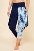 A person modeling loose harem-style cropped pants with a solid navy left leg and a blue-and-white tie-dye right leg, shown against a plain beige studio background.